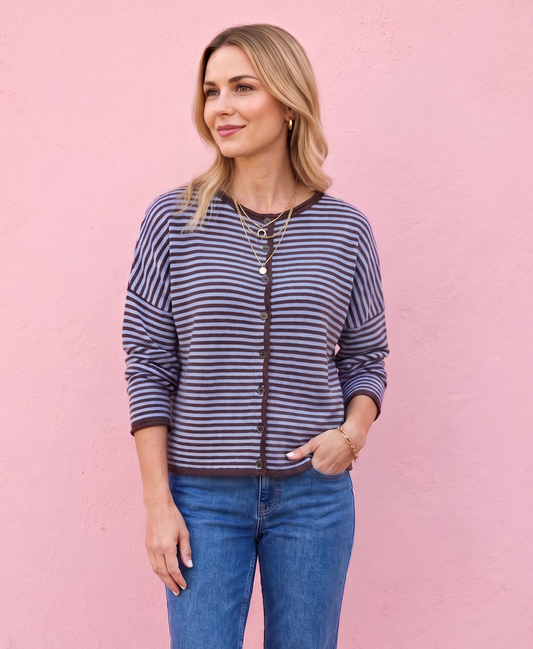 A woman stands against a pink wall wearing a navy and white striped zip-up cardigan, blue jeans, and dark brown boots.