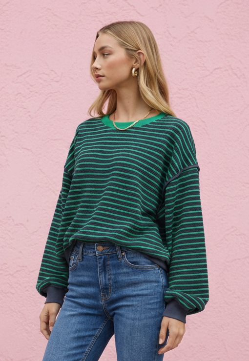 A woman stands against a pink textured wall wearing a green and dark green striped long-sleeve sweatshirt, blue jeans, and white sneakers.
