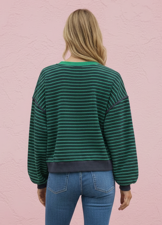 A woman viewed from behind wearing a green and black striped long-sleeve sweatshirt, blue jeans, and white sneakers, standing against a textured pink wall.