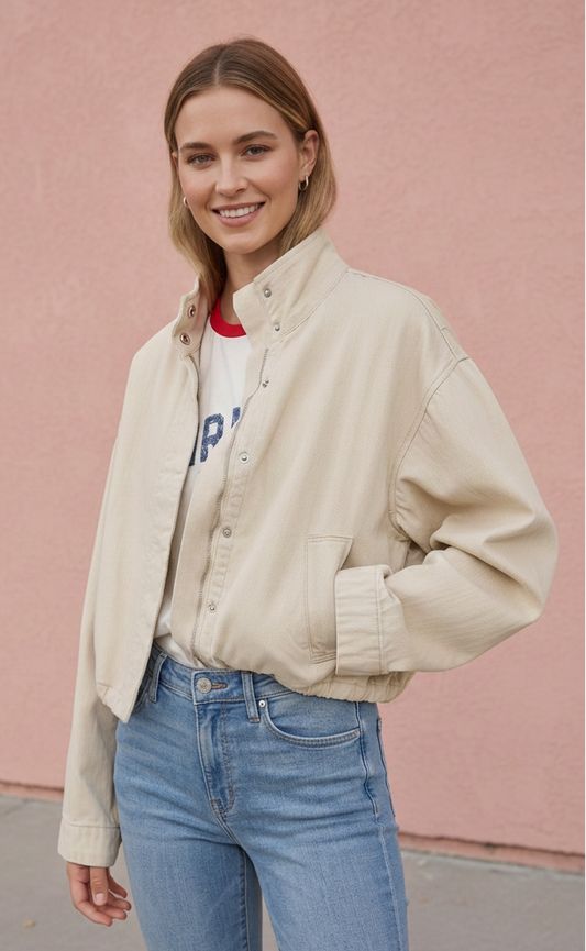 A woman wearing a light beige corduroy button-up jacket over a white graphic t-shirt and blue jeans, standing in front of a pink stucco wall.