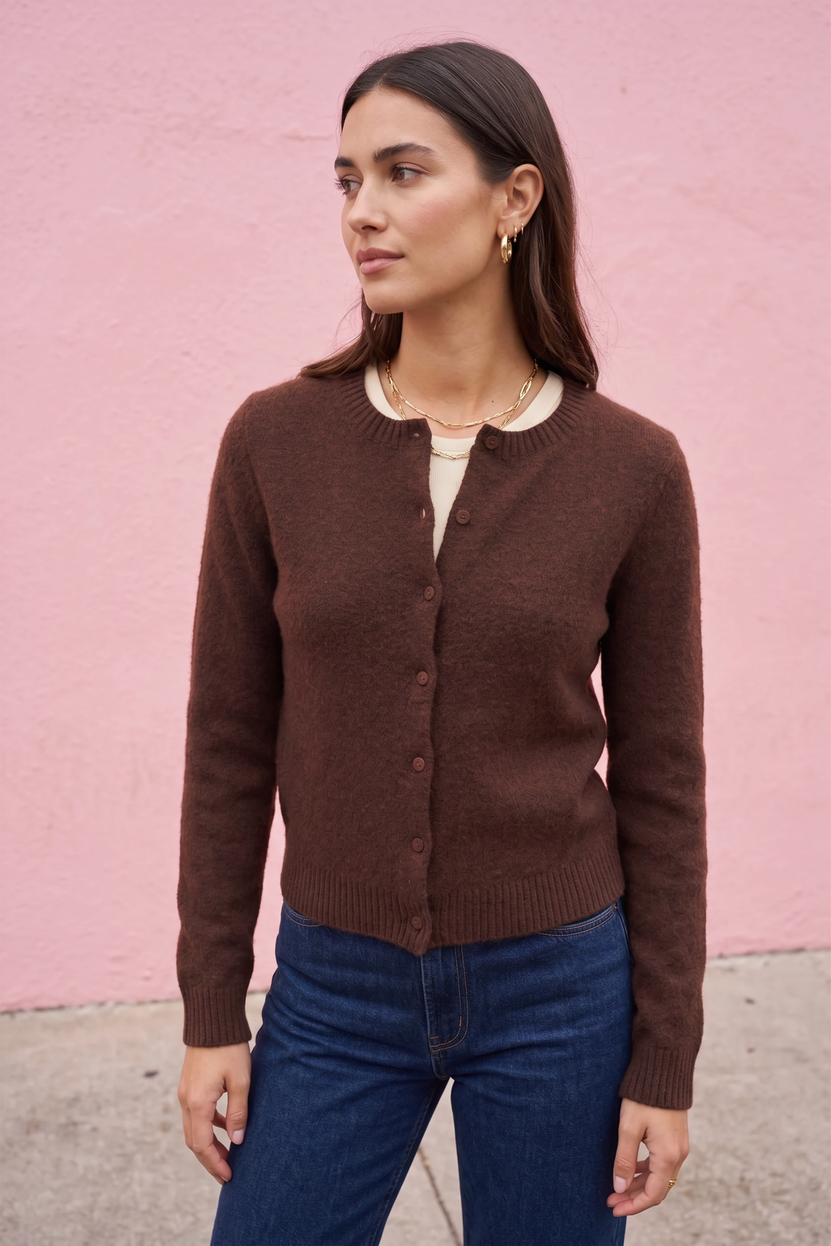 A woman wearing a brown button-front knit cardigan over a cream top and blue jeans, standing against a pink wall.