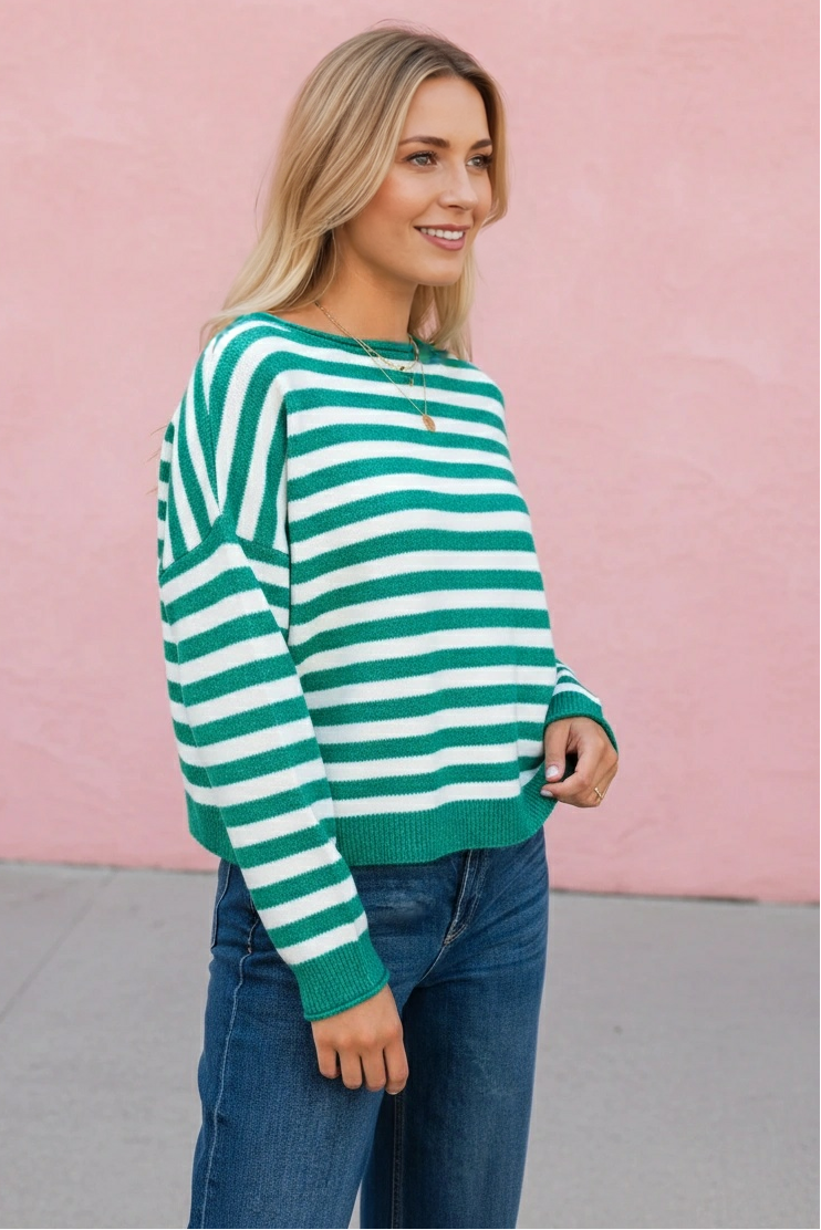 A woman wearing a green and white striped knit sweater and blue jeans, standing against a pink wall.