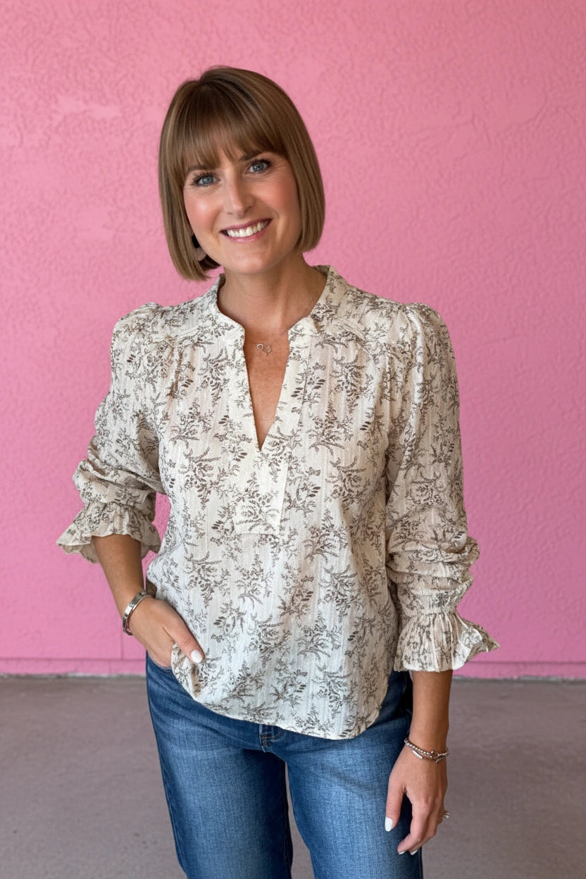 A woman with a short brown bob hairstyle wearing a cream-colored long-sleeve blouse with a brown floral pattern and blue jeans, standing against a textured pink wall.