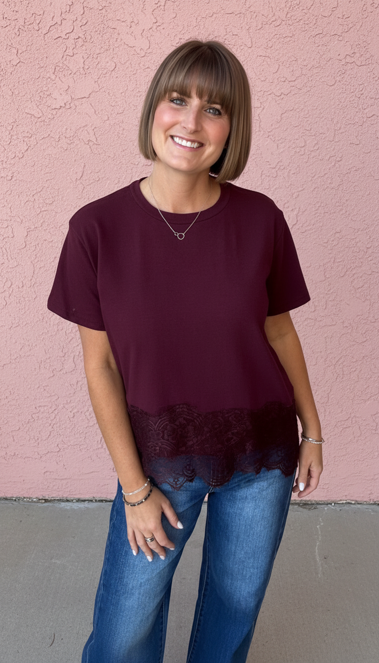 A woman with a short brown bob stands against a pink stucco wall wearing a short-sleeve crewneck t-shirt in deep burgundy with a black lace hem, paired with blue jeans.