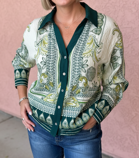 A woman with a short blonde bob stands against a pink stucco wall wearing a long-sleeve button-up shirt with a green and olive paisley pattern on an off-white base, paired with blue jeans.