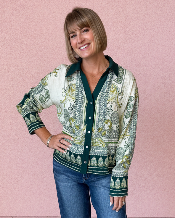 A woman stands barefoot against a pink wall wearing a long-sleeve button-front cardigan with a green and cream paisley pattern, paired with blue jeans.