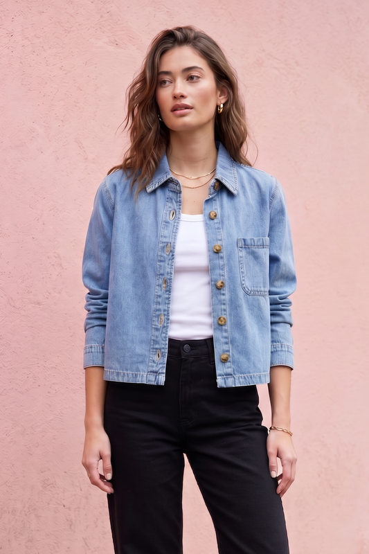 A woman stands outdoors against a pink stucco wall wearing a light blue denim jacket over a white top, black wide-leg pants, and black shoes.