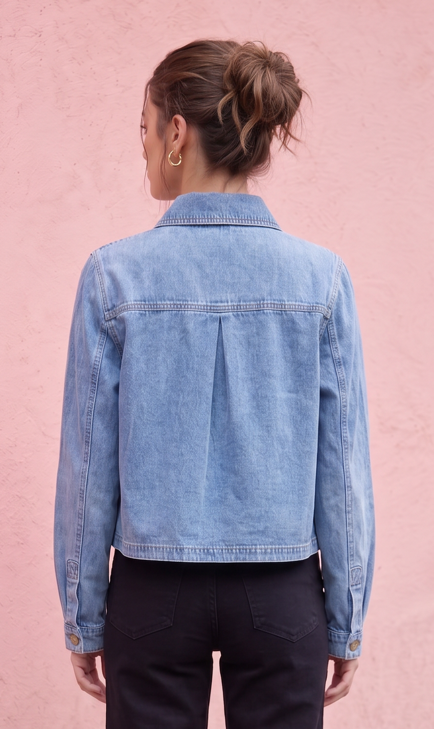 A woman stands with her back to the camera wearing a light blue denim jacket and dark charcoal pants, posing in front of a textured pink wall.