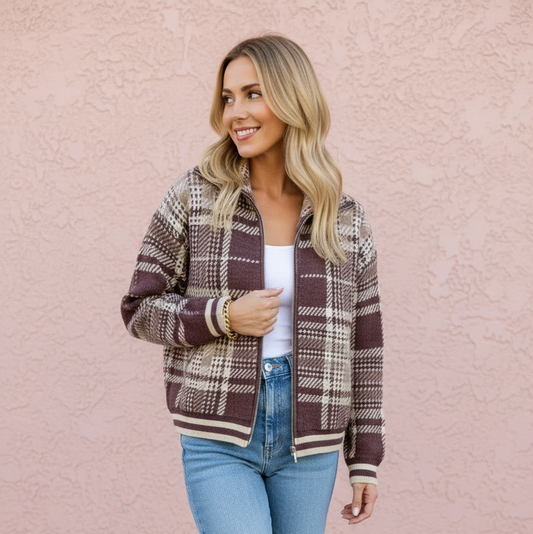 A woman wearing a brown and beige plaid zip-up jacket, a white top, and light blue jeans, standing against a textured pink wall.