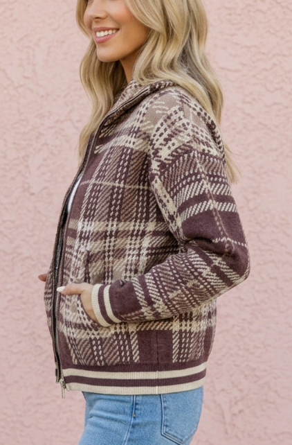A woman wearing a brown and beige plaid zip-up hoodie jacket with blue flare jeans, standing in profile against a textured pink stucco wall.