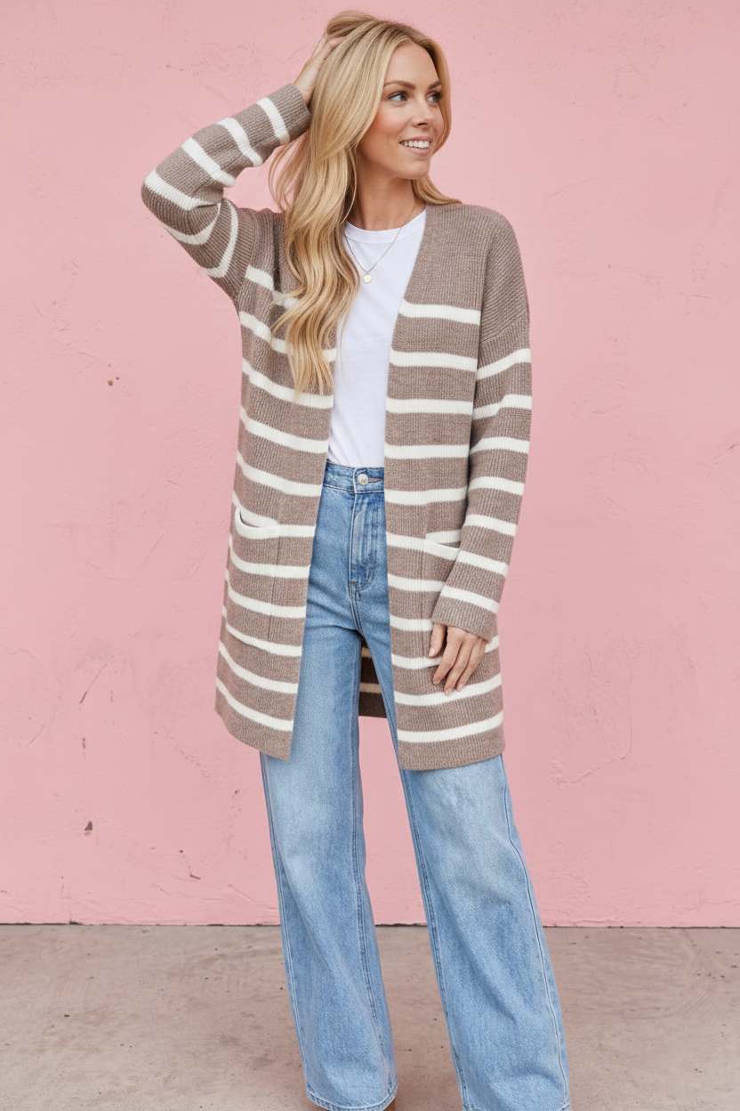 A woman stands against a pink wall wearing a long taupe and cream striped cardigan over a white top and light blue jeans.