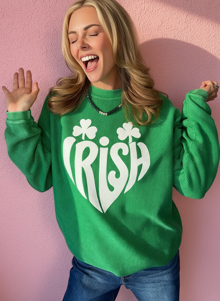 A woman wearing a bright green ribbed knit sweater with a large white heart and shamrock graphic spelling 'IRISH' on the front, posing against a pink wall.