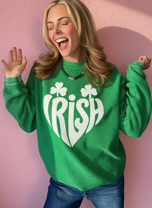A woman wearing a bright green ribbed knit sweater with a large white heart and shamrock graphic spelling 'IRISH' on the front, posing against a pink wall.