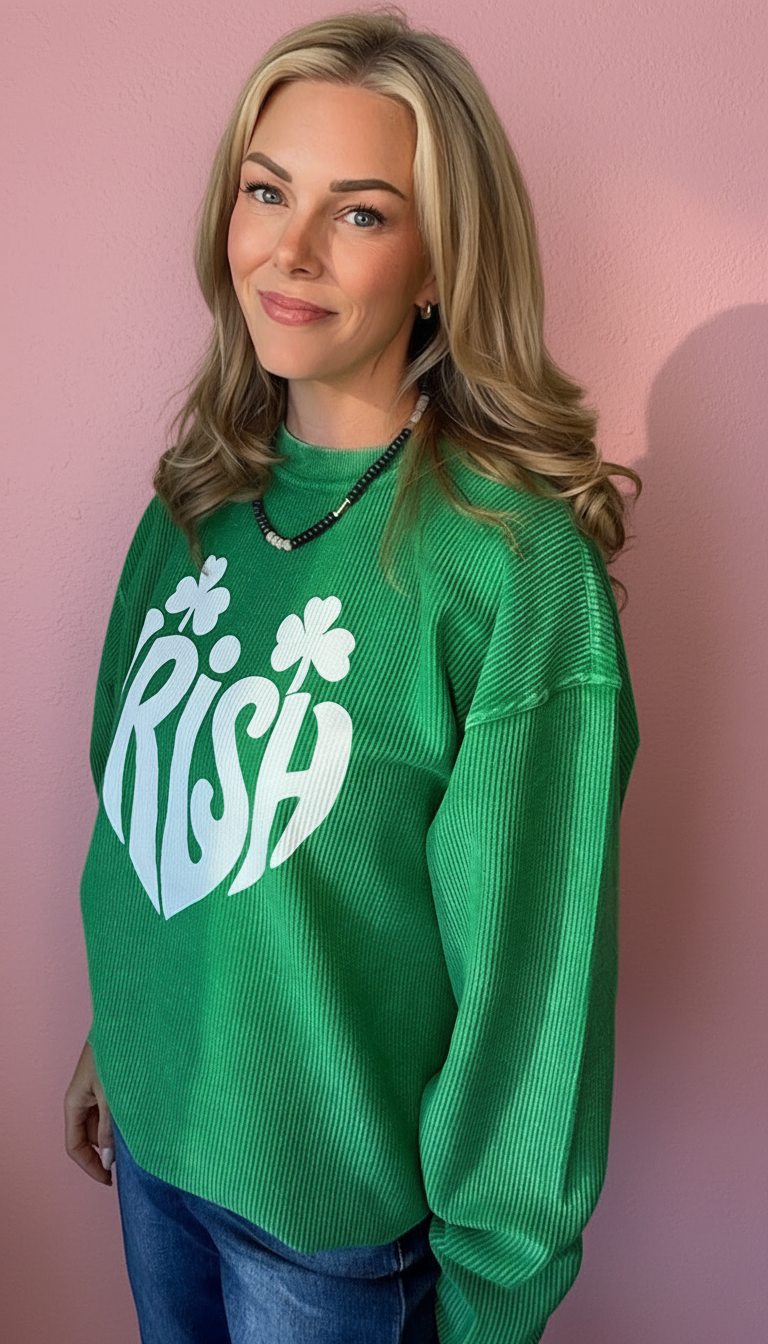 A woman with blonde hair wearing a bright green ribbed knit crewneck sweatshirt with a white 'Irish' shamrock graphic, standing against a pink wall.