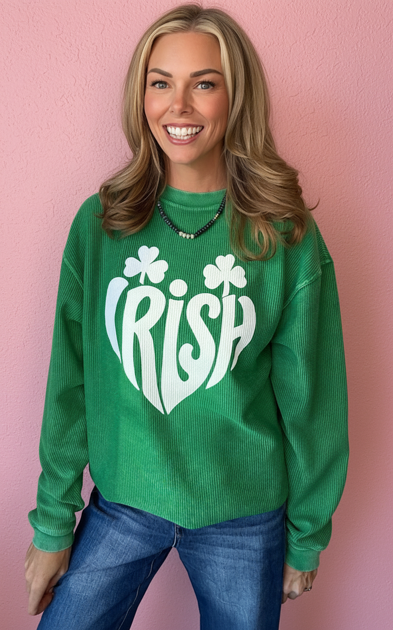 A smiling woman with blonde hair stands against a pink wall wearing a bright green ribbed sweatshirt with a large white 'IRISH' shamrock graphic on the front, paired with blue jeans.