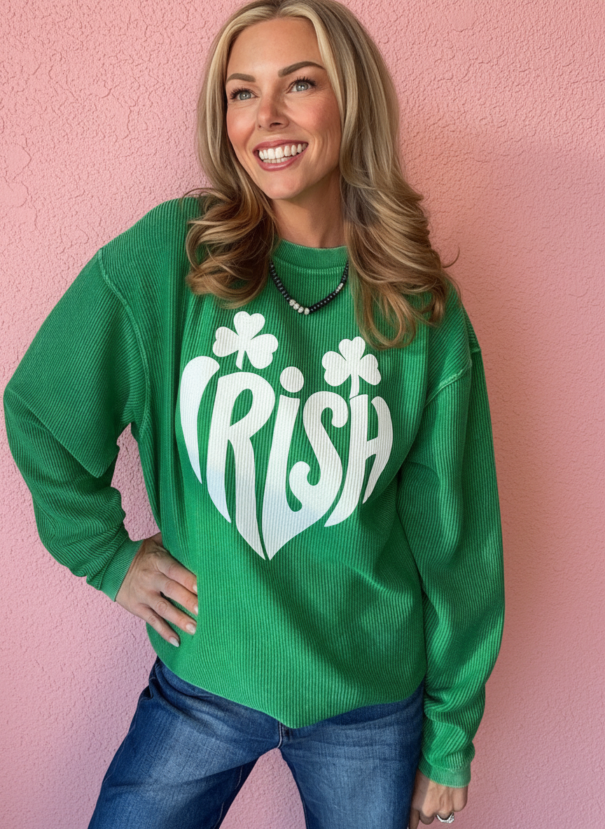 A woman wearing a bright green crewneck sweatshirt with a white graphic that reads 'IRISH' in a heart shape with shamrocks, paired with blue jeans, standing against a pink textured wall.