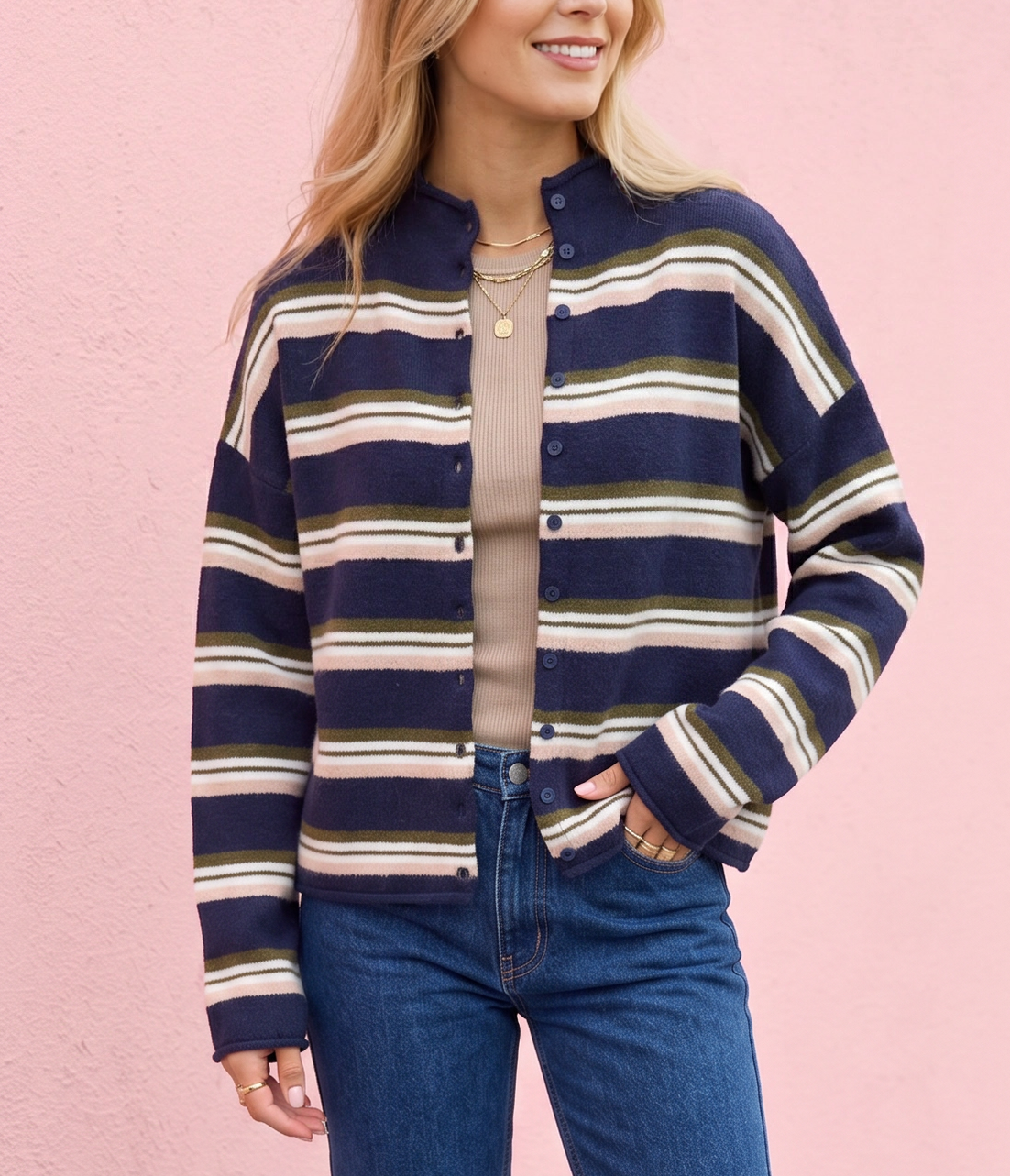 A woman stands against a pink wall wearing a navy, beige, and white striped zip-up jacket, blue jeans, and white sneakers.