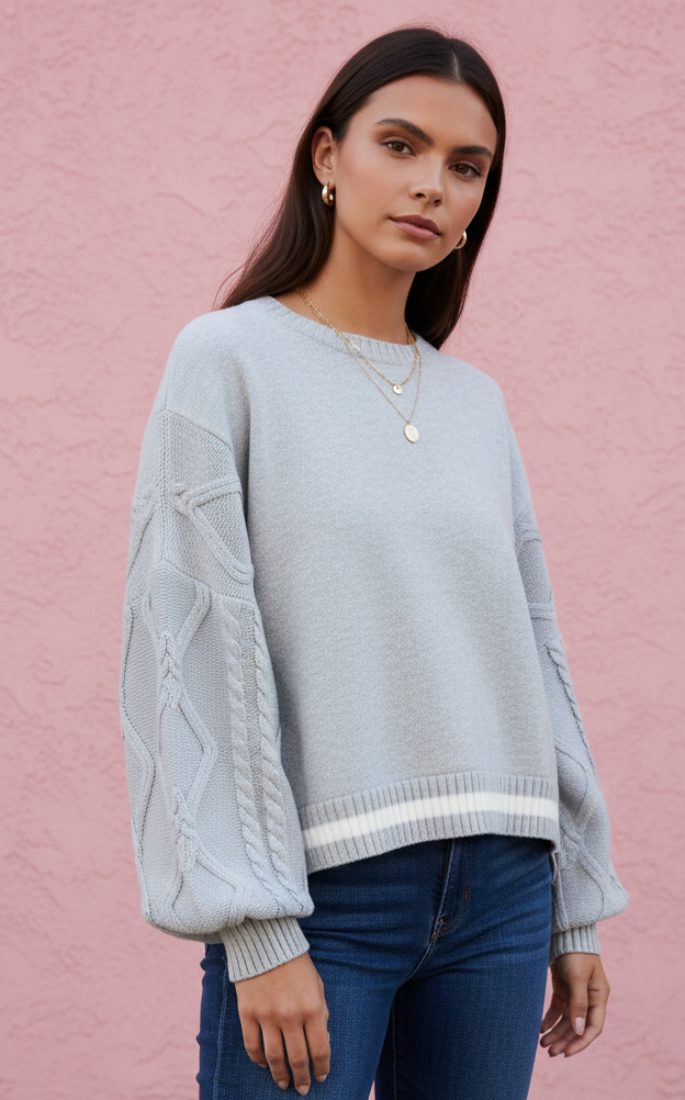 A woman wearing a light gray cable-knit sweater with balloon sleeves and blue jeans, standing against a textured pink wall.