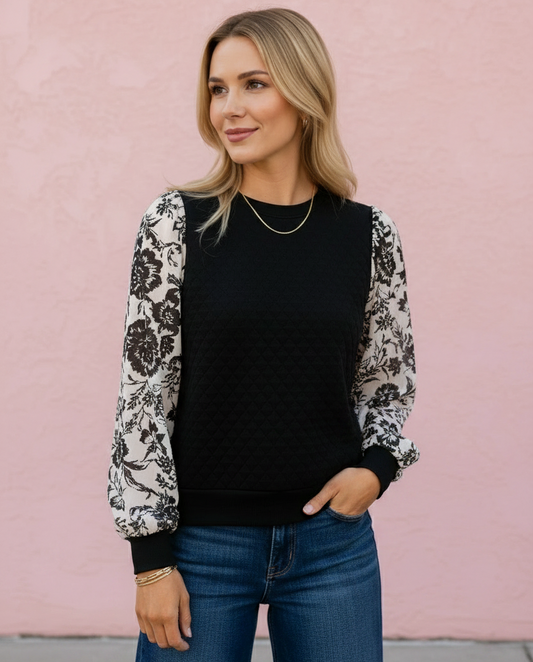 A woman wearing a black crewneck sweater with long black and white floral patterned sleeves, paired with blue jeans, standing against a textured pink wall.