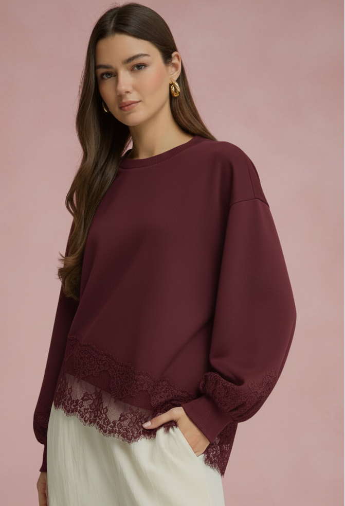 A woman modeling a long-sleeve crewneck top in deep burgundy with a lace hem and lace cuffs, paired with white wide-leg pants, set against a solid pink studio background.